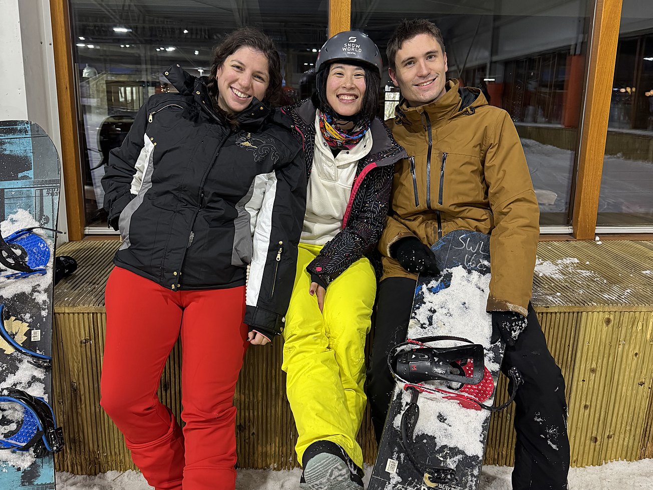 Eva, Malika et Mathis en tenue de ski, assis sur un banc enneigé devant les baies vitrées d'une piste couverte, planches de snowboard à la main