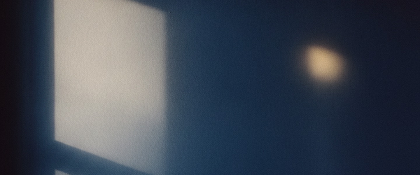 Mur bleu nuit mat en lumière rasante, traversé d'un trapèze de lumière et d'une lueur dorée